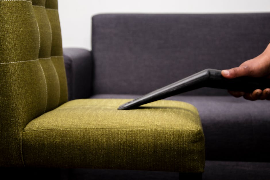A professional cleaner dressed in white protective gear, including a mask and gloves, is applying surface cleaning to a black leather office chair using an orange cloth in a well-lit modern living roo